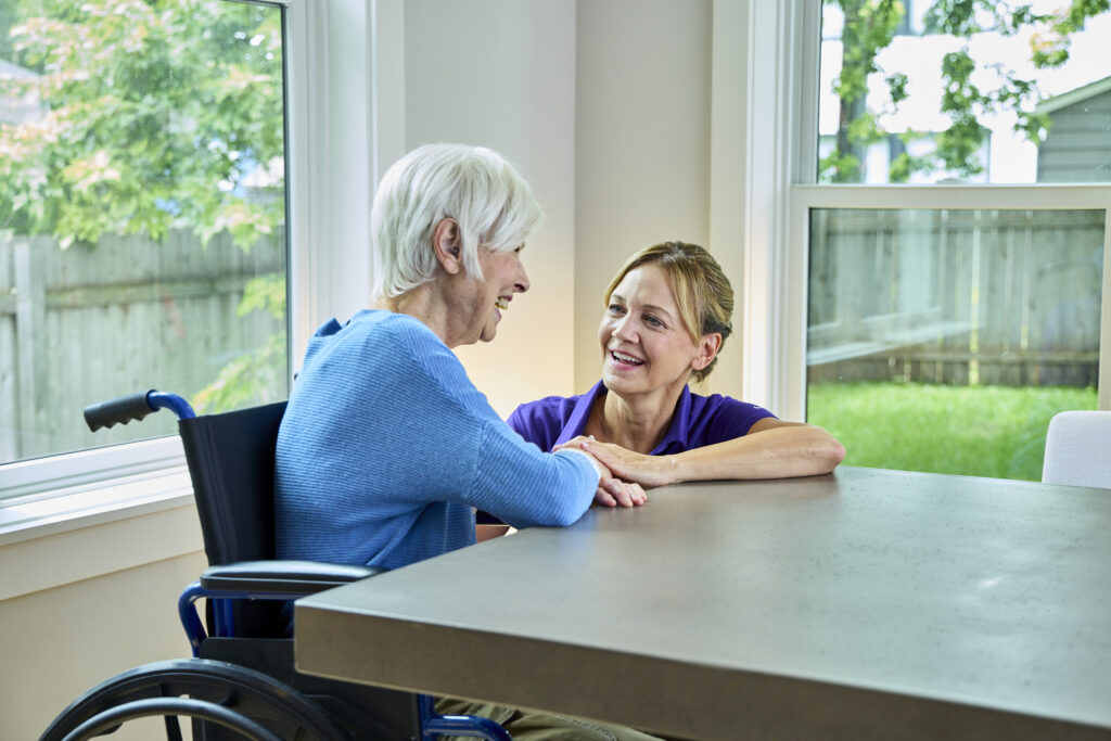 A smiling Touching Hearts at Home professional holding hands with a senior woman sitting in a wheelchair near a window, showcasing the personal touch driving senior care franchise profitability